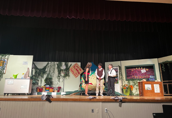A scene from the opening night of Little Shop of Horrors, with actors Jonah Gard, Sophia Batista, and Dylan Bond, playing characters Seymour, Audrey, and Mr. Mushnick, respectively.