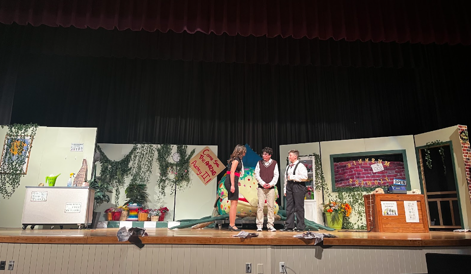 A scene from the opening night of Little Shop of Horrors, with actors Jonah Gard, Sophia Batista, and Dylan Bond, playing characters Seymour, Audrey, and Mr. Mushnick, respectively.

